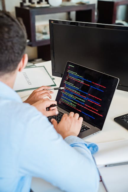 A person sitting at a desk using a laptop during a home relocation process, with the laptop displaying colorful lines of code indicating the loading or transfer of digital files. The workspace include